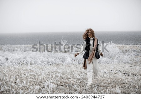 A happy red-haired girl in a white sweater and pants, a black vest and a long scarf is having fun in nature in winter. 