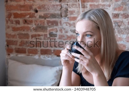 The close-up image captures a young woman with blonde hair as she takes a sip from a small coffee cup, her eyes looking off into the distance. She is sitting in a cozy corner of a cafe that boasts a