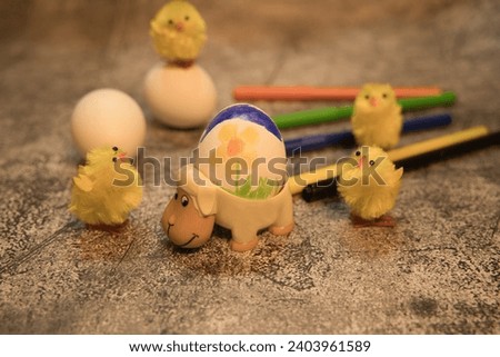 painted egg in an egg cup in the shape of a sheep with chick and pencils in the background