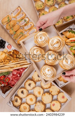 An overhead shot depicts a hand holding a box of lemon meringue tarts amidst a spread of assorted appetizers, suggesting a concept related to catering or party food