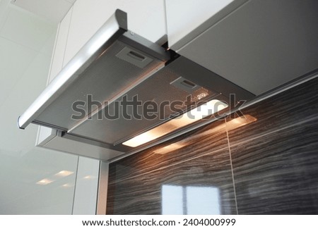 A close-up on a built in, integrated into kitchen cabinet stainless steel range hood, cooker hood, exhaust or extractor vent hood working with lights on in the kitchen. Royalty-Free Stock Photo #2404000999
