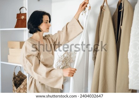 Young woman hold coat hanger with dress near wardrobe, choosing outfit for walk at modern apartment. Female neatly put apparel in closet, arranging clothes Royalty-Free Stock Photo #2404032647