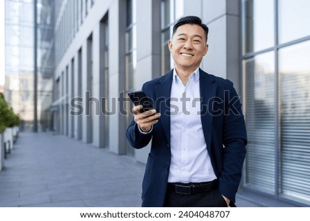 Portrait of a young Asian male businessman standing smiling on the street near an office center, holding a phone and looking at the camera. Royalty-Free Stock Photo #2404048707