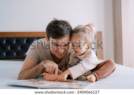 Dad and little girl are reading a book of fairy tales on the bed, running their fingers through the pages