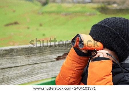 Boy in winter clothes looking the green meadow with binoculars during hiking day in the mountains
