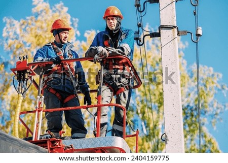 Two professional electricians in hard hats are repairing power lines from cradle of bucket truck. View from below. Electricians change cables on street lighting poles.