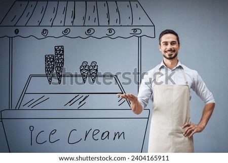 Handsome guy in apron is leaning on drawn ice cream cart, looking at camera and smiling while standing at the gray wall