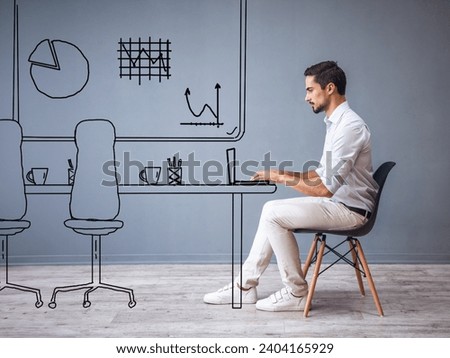 Handsome man in smart casual clothes is working with laptop while sitting on the chair on gray background with drawn office