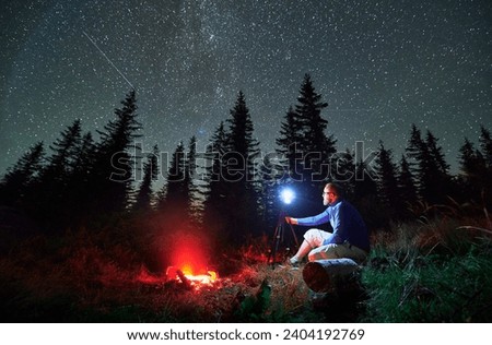 Male photographer doing photo shoot in mountains. Man taking pictures of starry night. Young man sitting on log next to bonfire at night.