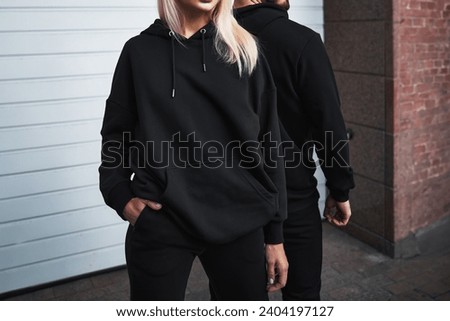A mock-up template of black hoodies worn by a couple. A design concept for print and branding. A couple poses outdoors in a casual and elegant street wear style. No face and no logo shown.