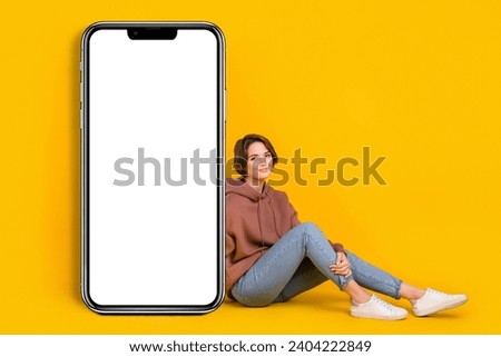 Full body photo of charming young lady sitting near smartphone touchscreen advert new web store page isolated on yellow color background