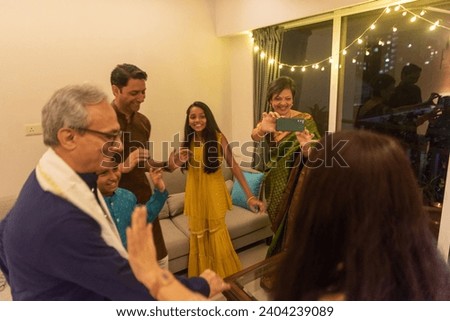 An Indian grandmother, wearing a woven green silk saree, takes a video with her cellphone of her daughter in law during an impromptu family dance party during dinner at their home in Mumbai, India. Royalty-Free Stock Photo #2404239089