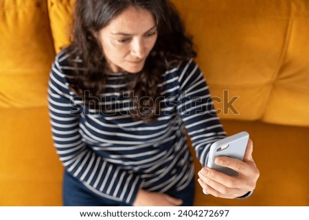 A middle-aged woman is sitting comfortably on a sofa, engaged in a video call on her smartphone. She appears relaxed and content as she communicates through technology.
