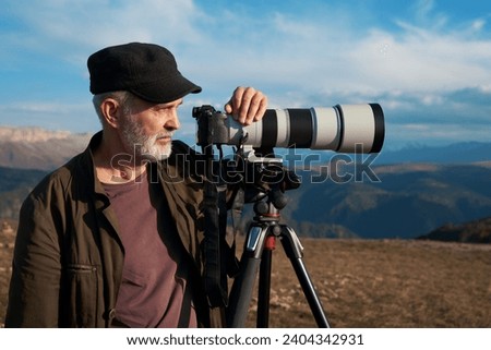 Photogaph (adult gray-haired man) with a camera with a long lens on a tripod on the background of mountainous terrain.                               