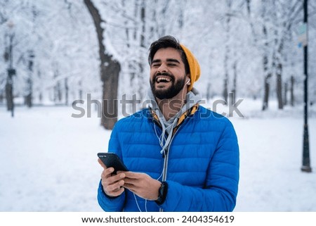 Content happy young man hearing good news over mobile phone smiling with eyes closed while standing in public park on snowy day in winter season.