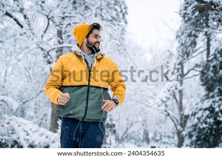 Joyful handsome male athlete wearing yellow beanie and weatherproof jacket running while listening to music in snowy forest during winter. Royalty-Free Stock Photo #2404354635