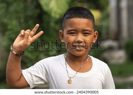 A young asian kid makes a peace sign. Wearing a white T-shirt. Outdoor garden scene.