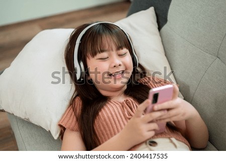 Young happy and adorable Asian girl in headphones is using a smartphone while lying on a sofa in the living room. Kid with modern technology concept