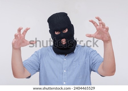 A scary and unhinged evil man wearing a black ski mask makes a threatening though comical gesture, showing his claws. Isolated on a white backdrop.