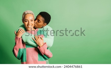 Two young cheerful women wearing green clothes embracing and kissing in a studio with a green background. Royalty-Free Stock Photo #2404476861