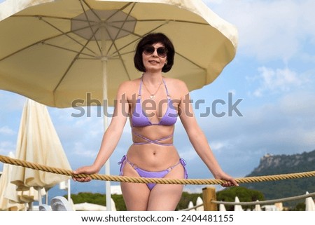 Woman 40-44 years old in a lilac bikini under a sun umbrella beach. Royalty-Free Stock Photo #2404481155