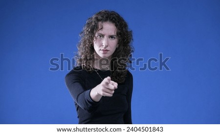 Woman accusing viewer by pointing finger at camera while standing on blue background. Upset doubtful person gesturing at offender Royalty-Free Stock Photo #2404501843