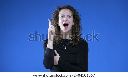 Thoughtful Woman with Sudden Insight, Eureka Epiphany on Blue background raising hand and finger in the air feeling enlightened Royalty-Free Stock Photo #2404501857