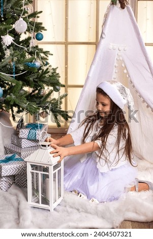 Happy kid girl 5 year old with lamp posing near Christmas tree, smiling look. Adorable child girl in white dress and santa hat at wigwam in children room. Christmas magic concept. Copy ad text space