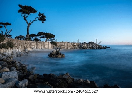 Dramatic long exposure image in early morning sunrise of the pacific coast in Monterey, California at lover’s point in Pacific grove, with blurred bay and silhouette trees in background.