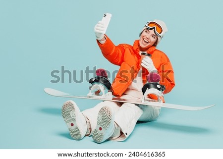 Full body young woman in warm windbreaker jacket ski goggles mask sit with snowboard do selfie mobile cell phone travel rest spend weekend winter season in mountains isolated on plain blue background