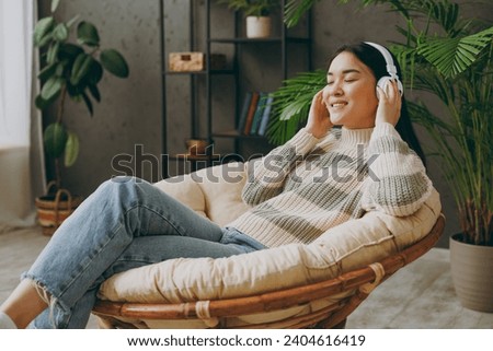 Side view young woman she wear casual clothes listen to music in headphones sits on armchair stay at home hotel flat rest relax spend free spare time in grey living room indoor. People lounge concept