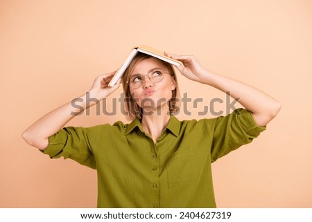 Photo of confused dissatisfied woman dressed green shirt hold book on head look empty space pouted lips isolated on beige color background