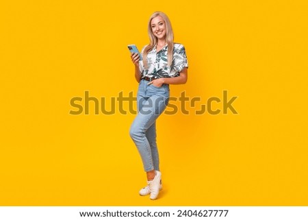 Full size photo of optimistic girl dressed print shirt stylish jeans holding smartphone arm in pocket isolated on yellow color background