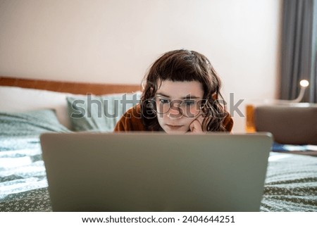 Intrigued concentrated serious teenager girl spending weekend, pastime at home, lying on bed, watching interesting film, video, movie on laptop computer, relaxing, reading online book attentively  Royalty-Free Stock Photo #2404644251