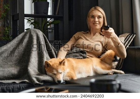 Young smiling blonde lying on couch at home watching television with her dog