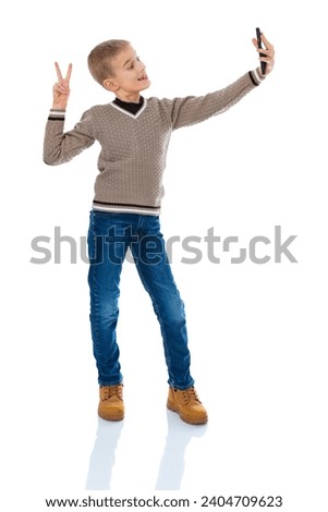 Full-length portrait of blonde seven year old boy take selfie and make v-signs isolated on white background. Blonde school boy in blue jeans holding cell phone and having video call posing in studio.