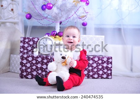 Little boy in Santa suit playing with a gift box under christmas tree in wooden Xmas decorated interior. Little boy holding present box in Christmas interior at home.