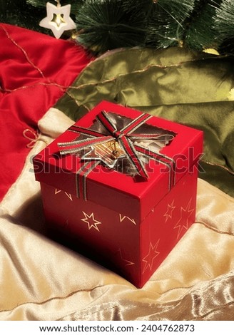 A red-boxed gift with a ribbon sits under the Christmas tree, serving as a New Year's present. It is adorned with Christmas decorations in a vertical photograph
