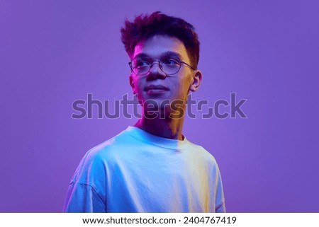 Portrait of smiling young man, boy in casual white t-shirt and sunglasses looking away against purple background in neon light. Relaxed look. Concept of human emotions, youth, education, lifestyle, ad Royalty-Free Stock Photo #2404767419
