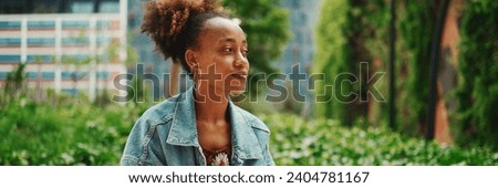 African girl with ponytail wearing denim jacket leaving voice message on mobile phone against modern city background.