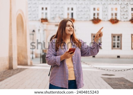 Picture of pretty young woman with backpack staying on the street. 30s tourist walking with phone and pointing finger on old city street. Use technology concept, Traveling Europe in summer