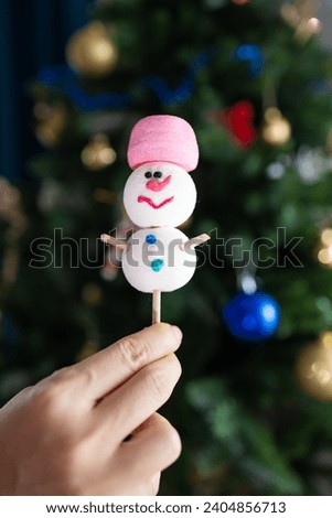 Hand holding marshmallow snowman near Christmas tree.