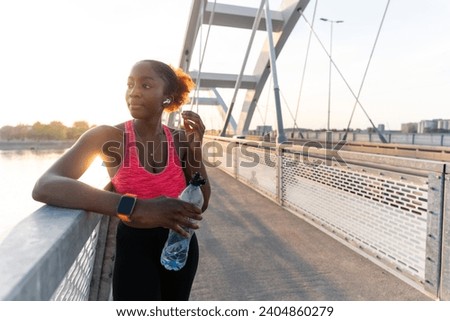Standing on the bridge at sunset, the African American girl leans against the railing, holding a bottle in her hand.