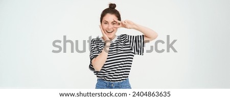 Beautiful brunette woman shows peace sign near eye and smiling, isolated against white background.