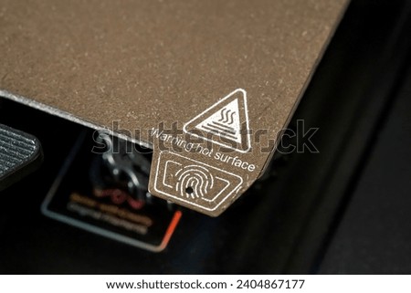 A 3D printer heated bed building pad hot surface warning label seen from up close, nobody. 3D printing equipment and accessories simple concept, device usage instructions warning icons symbols detail Royalty-Free Stock Photo #2404867177