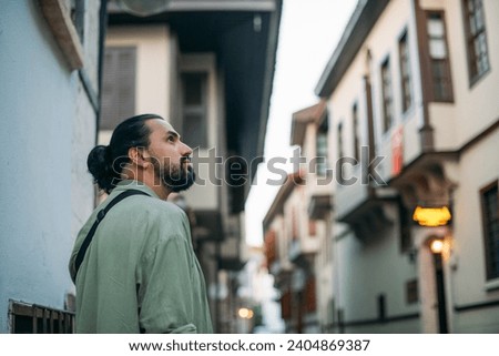 Portrait of a male tourist on a picturesque street of the old town.  A young, handsome guy in a light shirt walks through the green, narrow streets of the ancient city center Royalty-Free Stock Photo #2404869387