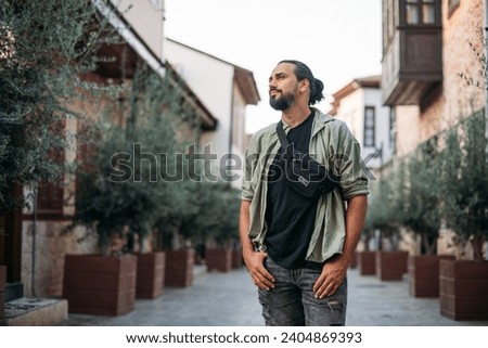 Portrait of a male tourist on a picturesque street of the old town.  A young, handsome guy in a light shirt walks through the green, narrow streets of the ancient city center Royalty-Free Stock Photo #2404869393