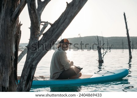 A man is surfing on a sup board on calm water at sunset. A young guy swims on a surfboard on a mountain lake among ancient, dry trees in the setting sun