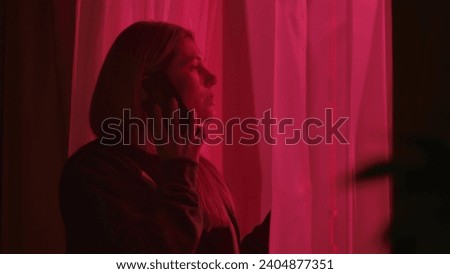 A woman standing near a window in a dark room illuminated by police lights, close up. Concerned woman talking on a smartphone.