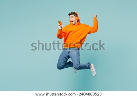 Full body side view young man he wearing orange hoody casual clothes jump high use hold mobile cell phone listen to music in headphones isolated on plain blue cyan color background. Lifestyle concept